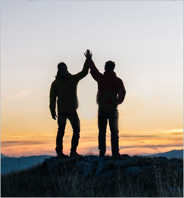 Two people on top of a mountain, high fiving in front of the sunset